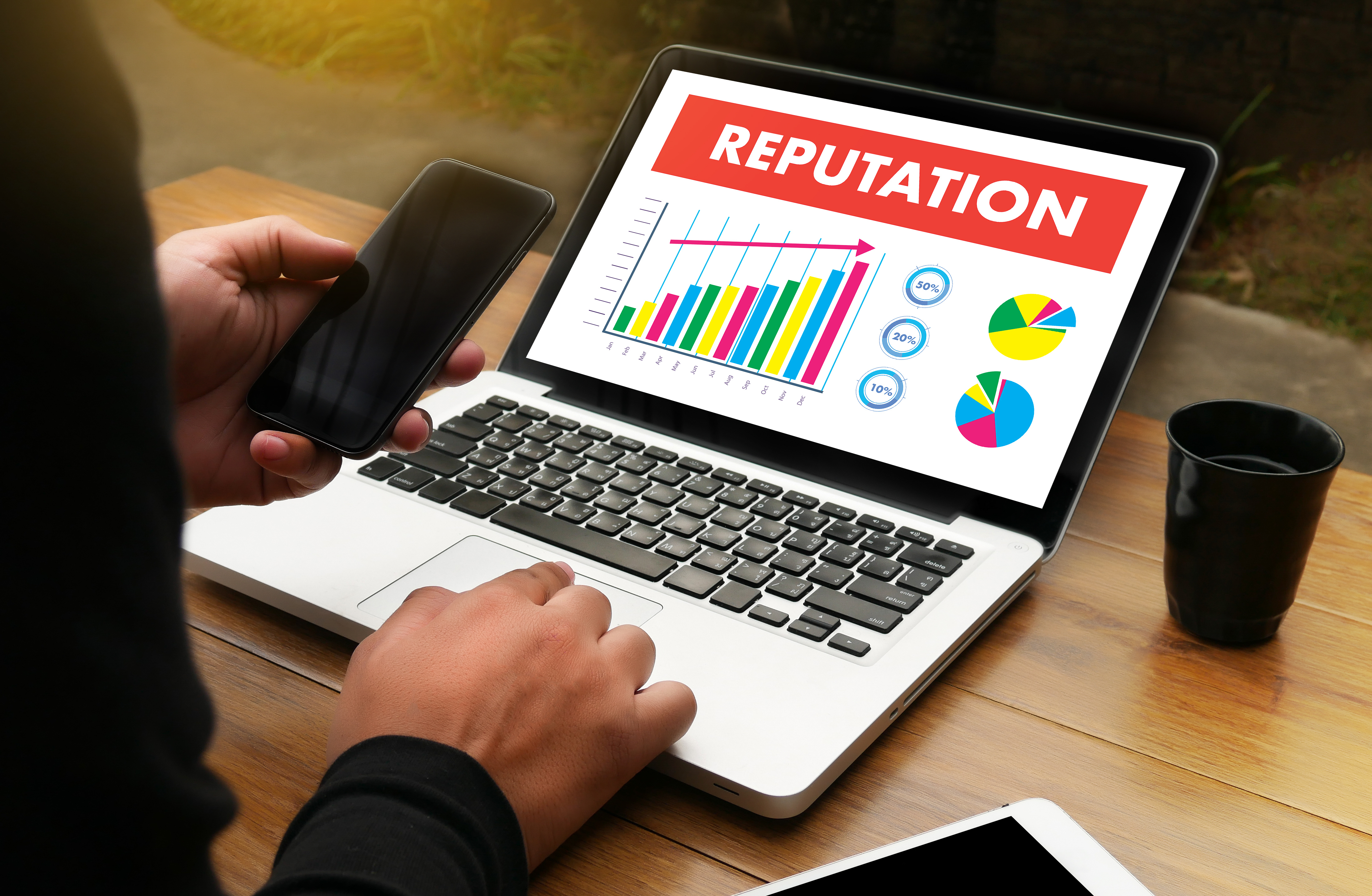 Somebody is sat in front of their laptop with their mobilephone in one hand and 
                using their laptop with the other. There is a dashboard on the screen called REPUTATION with some charts and graphs
