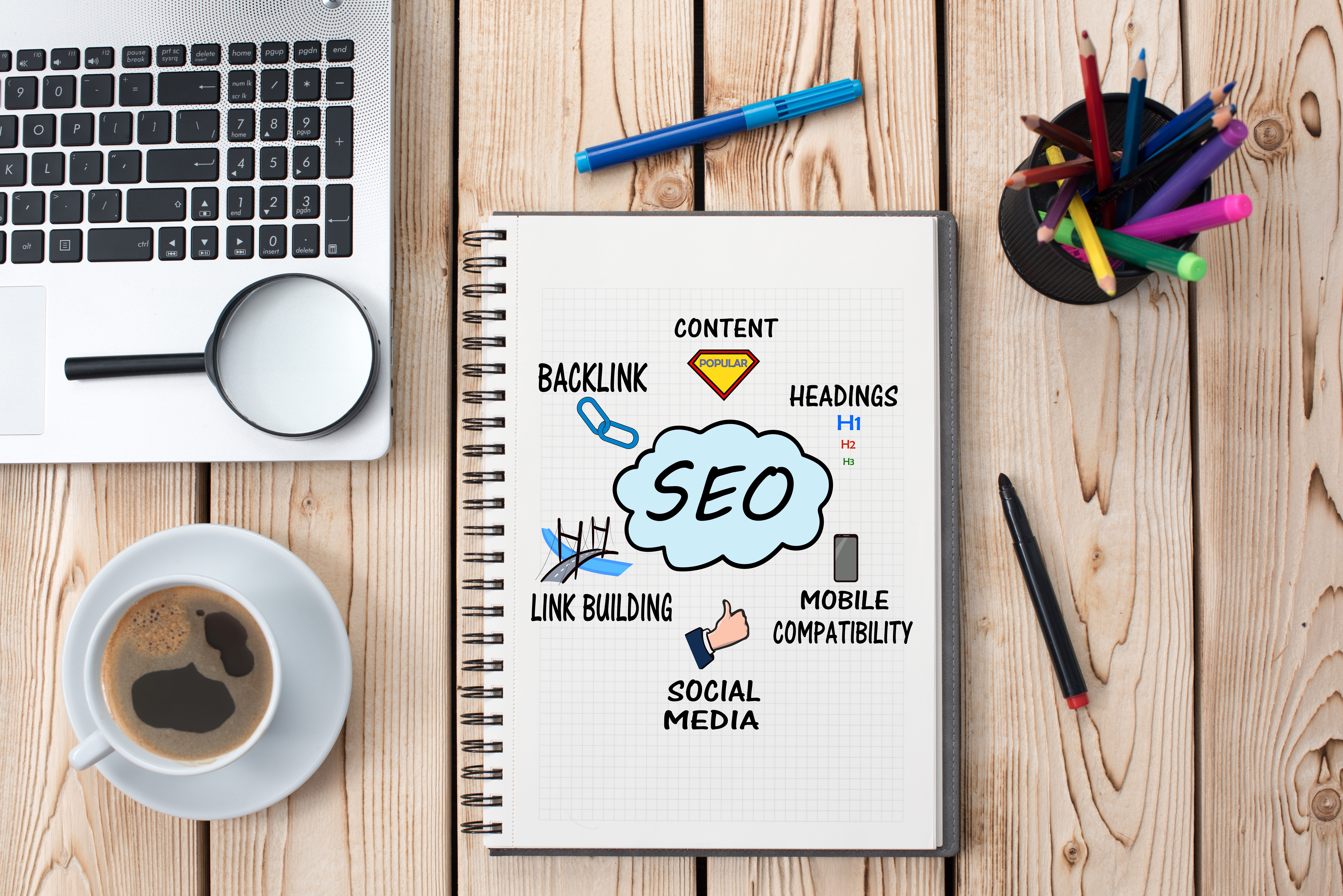 A notebook containing SEO notes sits on a desk next to a laptop and a cup of coffee. 
                The front cover contains headings such as backlink, content, headings, social media and link building